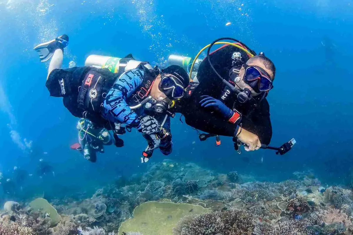 Two divers in full kit finning over a coral reef in clear blue water
