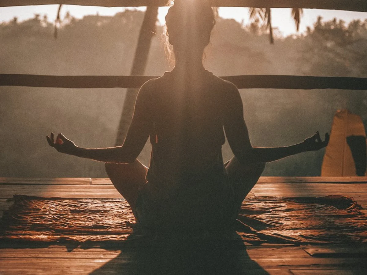 A person seated cross-legged in meditation on a timber deck, backlit by a low sun