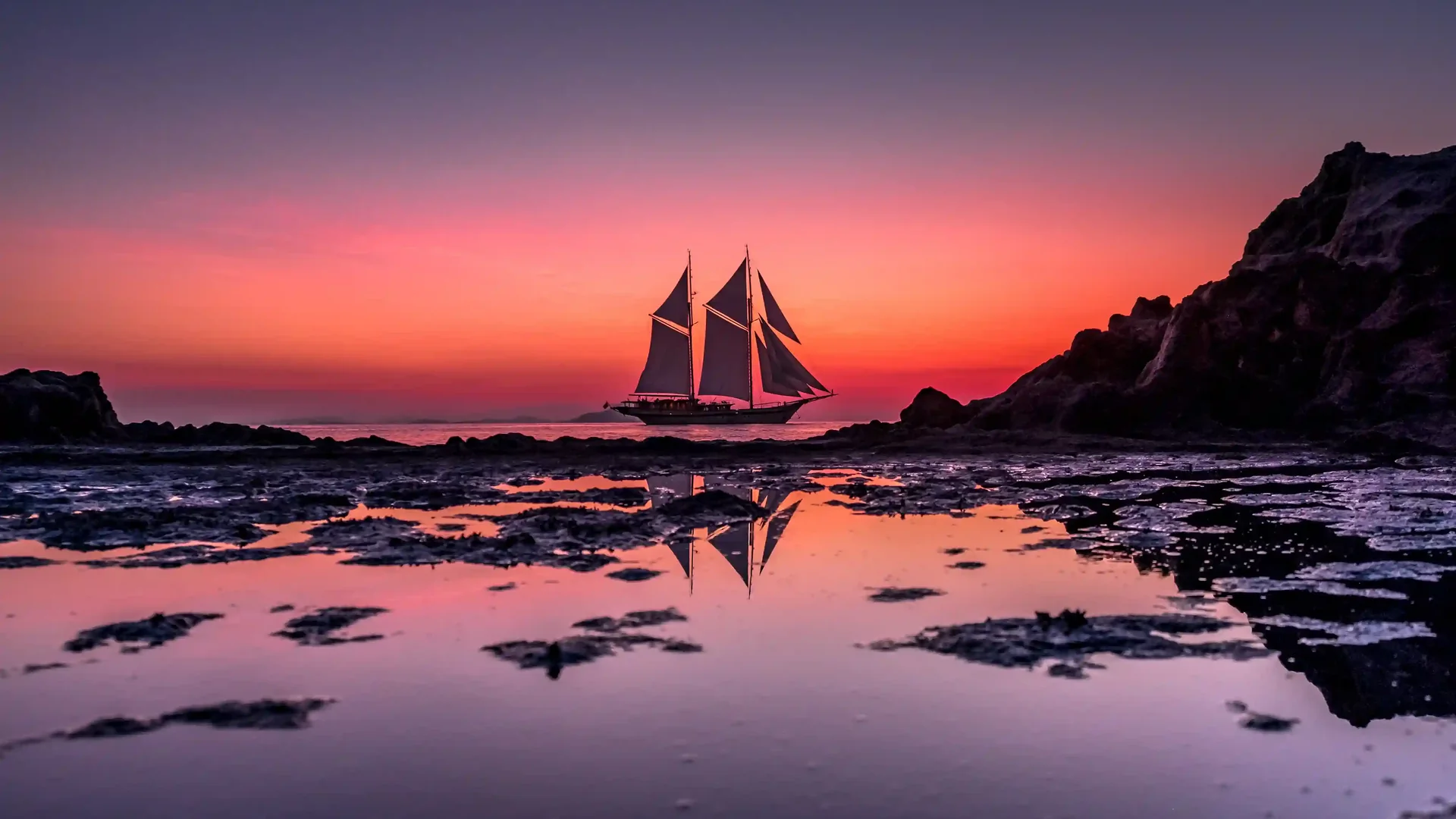 A phinisi under sail silhouetted against a deep red sunset over the Flores Sea