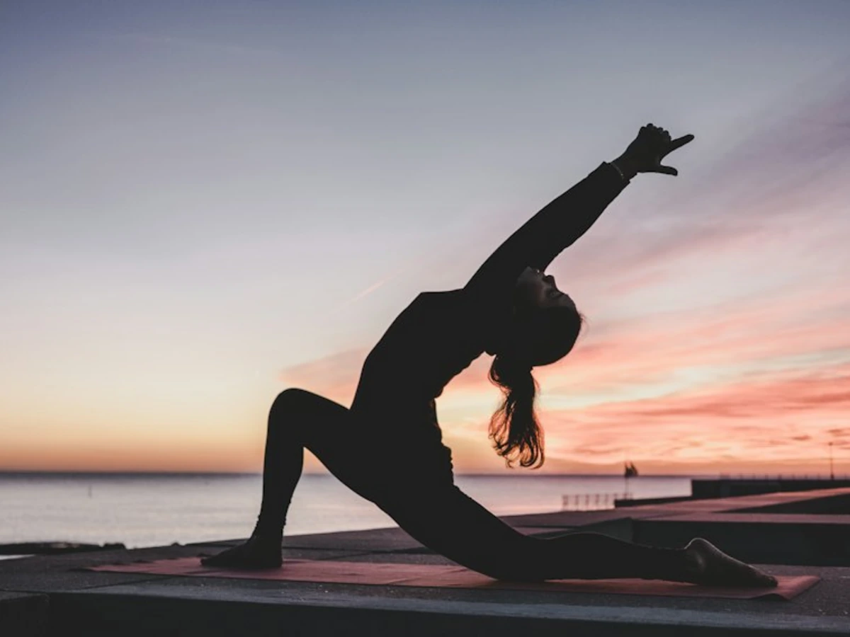 A yoga practitioner in silhouette holding a low lunge on a deck above water at sunrise
