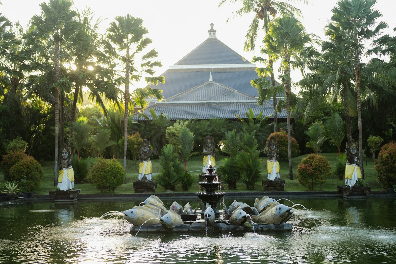 Fountain in a lush garden with palm trees and building.